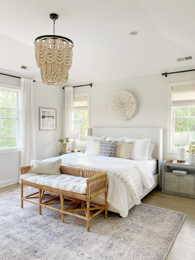 Neutral bedroom design features Loloi loren rug in slate, Wayfair upholstered bed in zuma white, rattan bench, beaded chandelier, and Benjamin Moore classic gray walls at 50% strength.