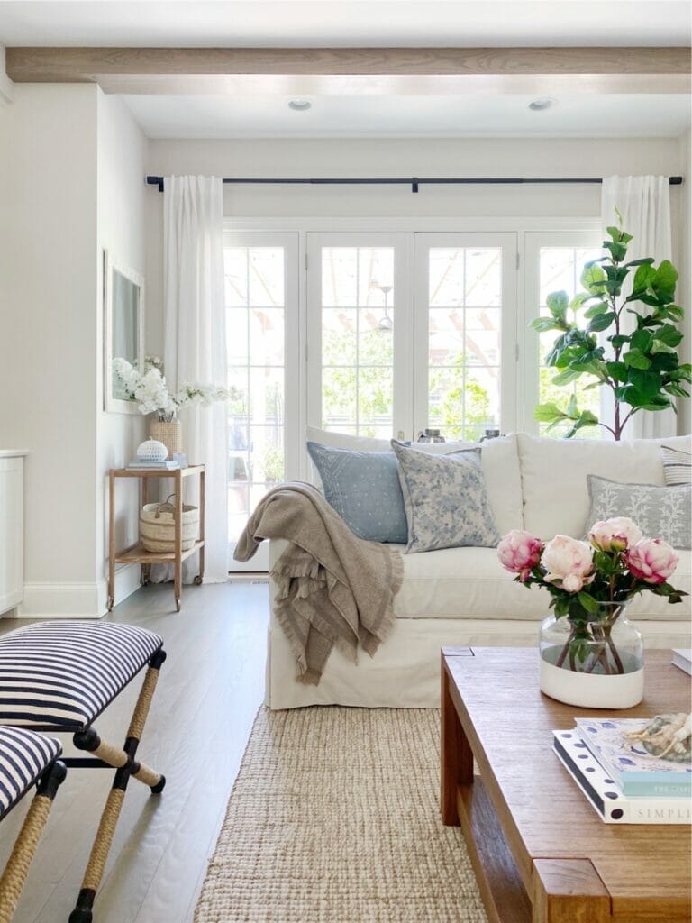Light filled spring living room with white slipcovered sofas, wool jute rug, striped stools from Wayfair, linen curtains, and pretty faux fiddle.