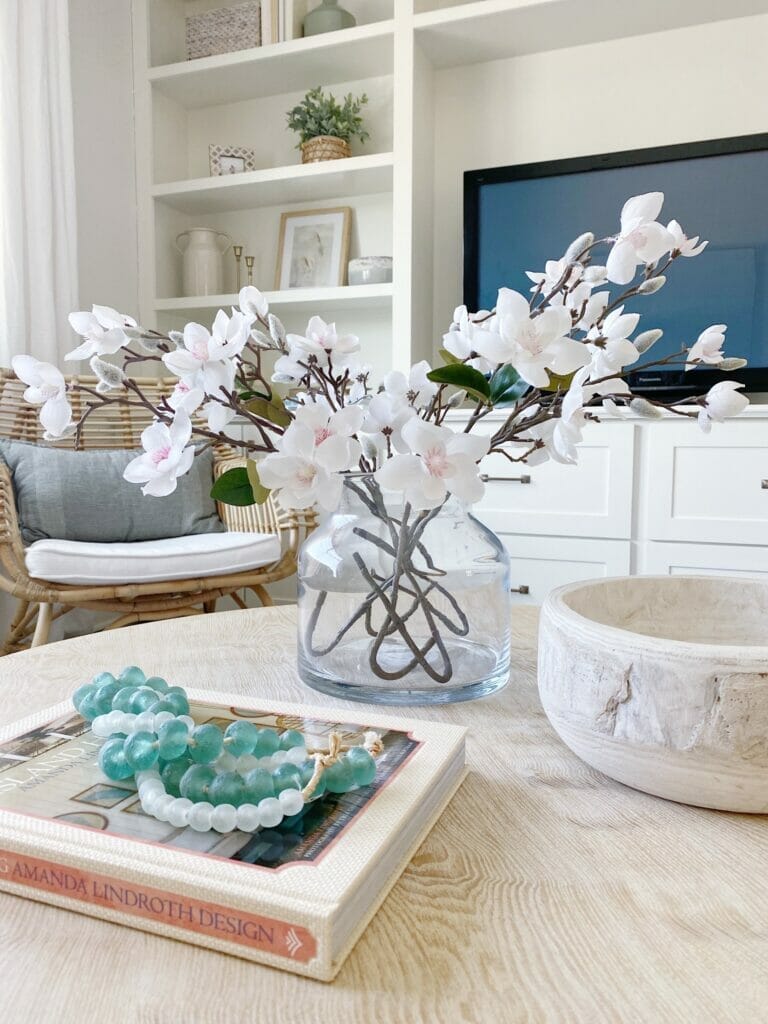 Coffee table styling - glass vase with faux stems, wooden bowl, and sea hued beads on book. 