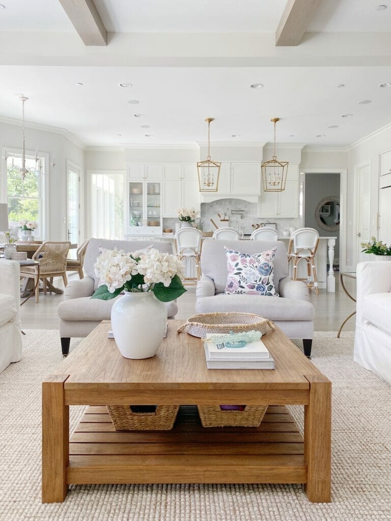 Memorial Day Sales Roundup - Summer living room with square wood coffee table, light grey upholstered chairs, open to kitchen with brass lanterns and white Serena & Lily riviera stools. Walls are benjamin moore classic gray. 