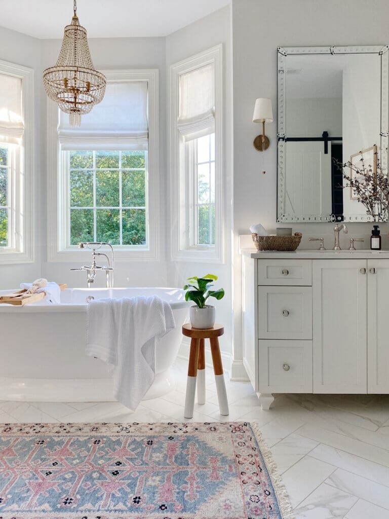 Primary bathroom features freestanding bath tub, vintage inspired rug, white vanity with large mirror and brass sconces
