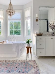 Primary bathroom features freestanding bath tub, vintage inspired rug, white vanity with large mirror and brass sconces
